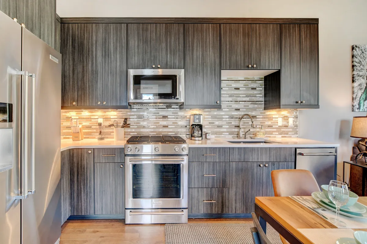 A bright, modern kitchen with gleaming stainless steel appliances, a custom decorative backsplash, and rich wooden cabinetry. The scene includes a set dining table, blending the kitchen and dining areas, and suggesting a space designed for both culinary creation and social connection.