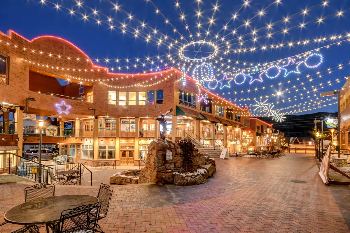 A bustling downtown scene featuring string lights overhead, decorative storefronts, and a central fountain. The area is lively and festive, ideal for shopping, dining, or grabbing ice cream with the kids.