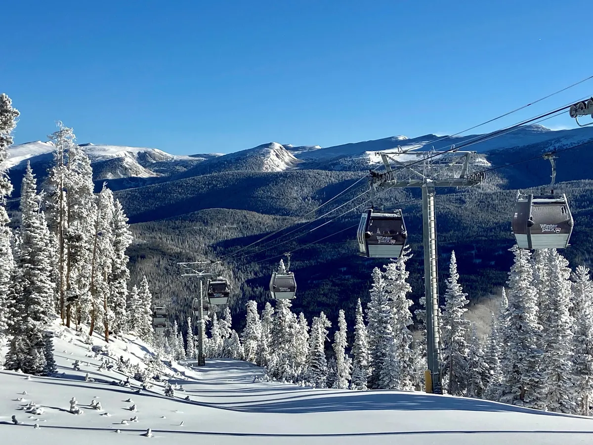 An exterior winter scene featuring gondolas ascending over snow-laden trees with majestic mountain peaks in the background. The scene evokes the excitement of skiing and snowboarding, with an emphasis on the proximity to world-class slopes.