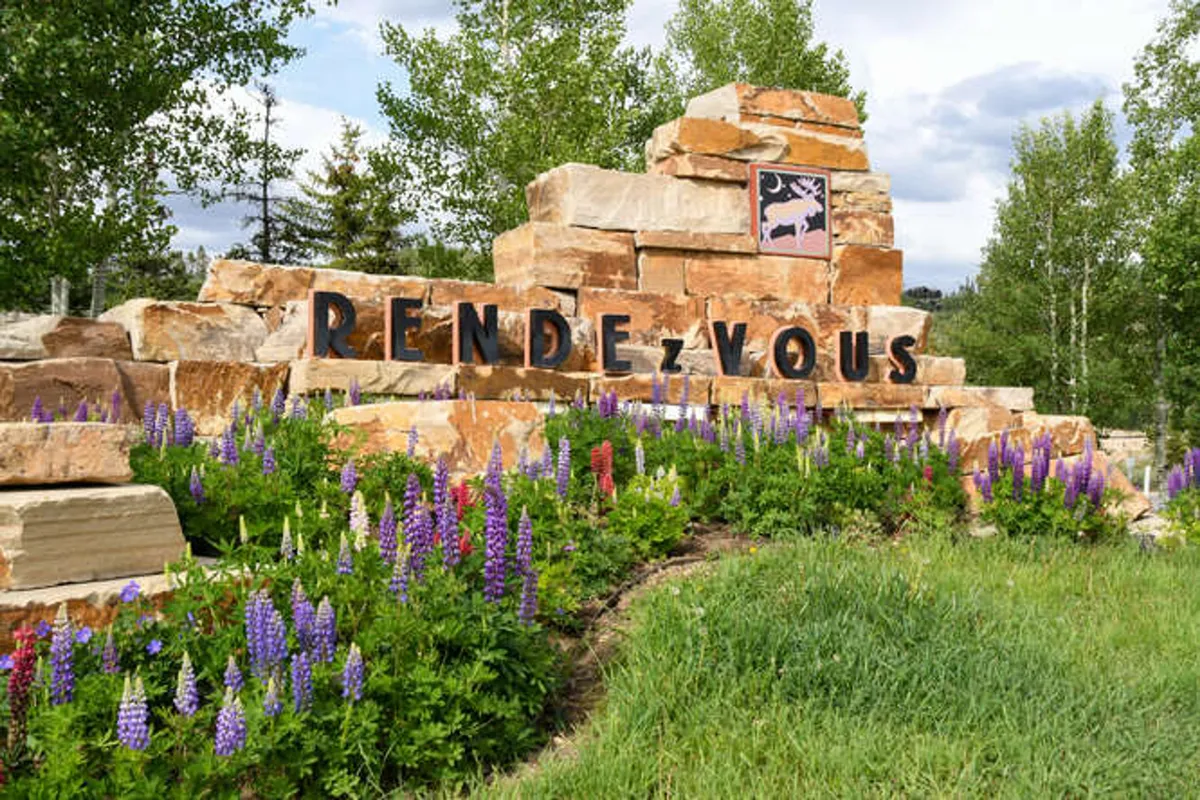 A welcoming exterior shot featuring clear signage, vibrant flower beds, and a charming stone structure set against a natural mountain backdrop. The scene conveys a sense of arrival and the scenic beauty of the community.