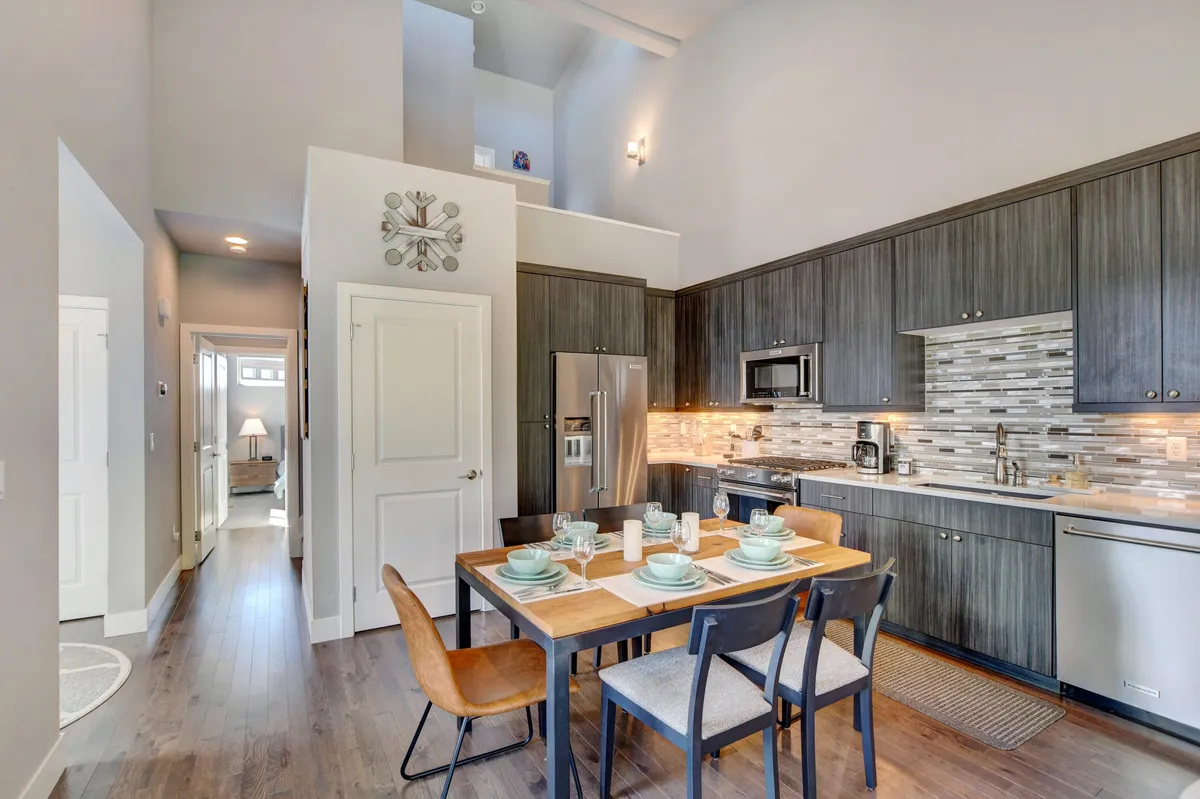 A welcoming interior view highlighting a large dining table set for a meal, with the modern kitchen and its appliances, textured backsplash, and warm lighting visible in the background. The ambiance is cozy, inviting, and perfect for gatherings.