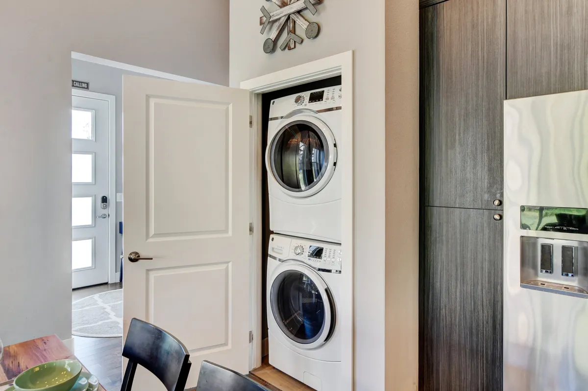 The image shows a stacked washer and dryer, a modern clock, and an open door leading to the exterior. The practical mood and visible access to the outdoors underscore the ease of preparing for spontaneous outdoor activities, such as a quick hike or bike ride.