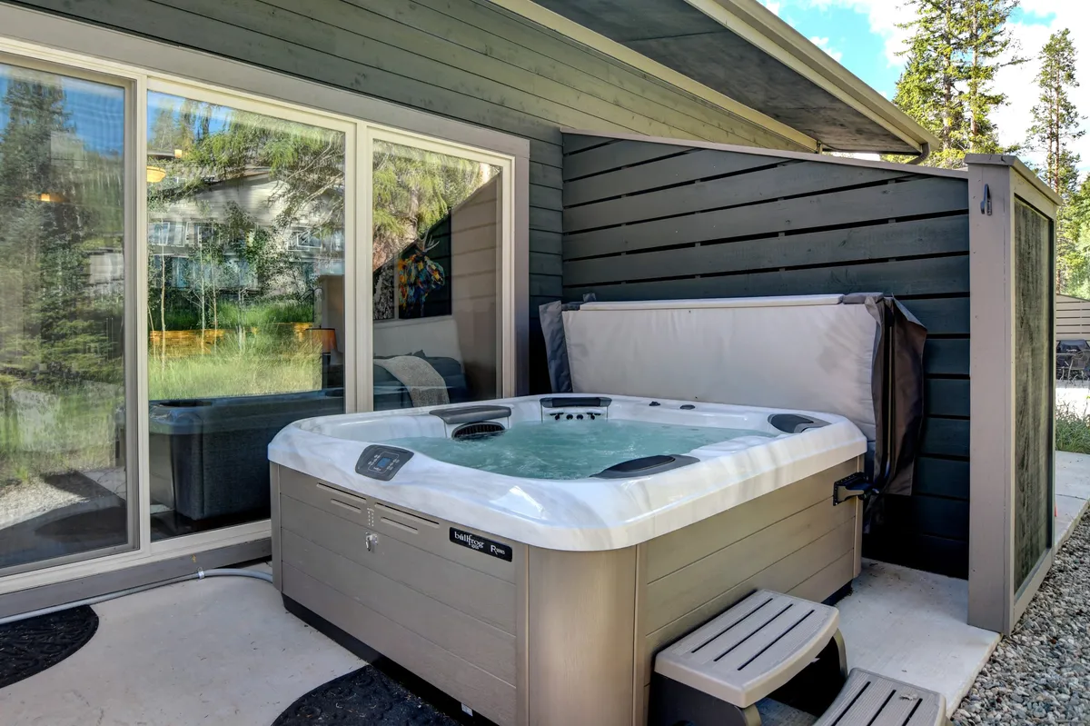 This image features a tranquil outdoor scene with a private hot tub positioned near glass sliding doors that open into the home, and a warm wooden wall providing a cozy backdrop. There are hints of surrounding nature, inviting guests to relax and soak in the views right from the patio space.
